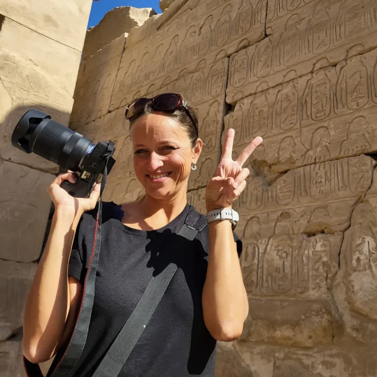 mladá usmívající se žena s profesionálním fotoaparátem a prsty zdviženými ve výtězném gestu V, na poozadí egyptských památek s hieroglify jako autor blogu pastelkomat.cz